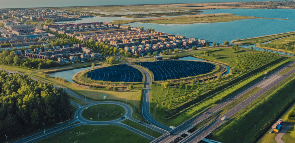Drone shot waarbij je een snelweg ziet, een woonwijk aan het water en een groot veld met zonnepanelen.