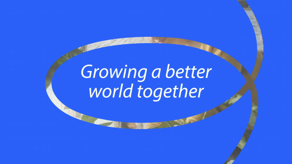 Video van een vloeiende grafische lijn over een blauwe achtergrond met daarop de tekst ‘Growing a better world together’. De lijn maakt een overgang in beeld naar een video van een vrouw die in een kas aan het werken is.